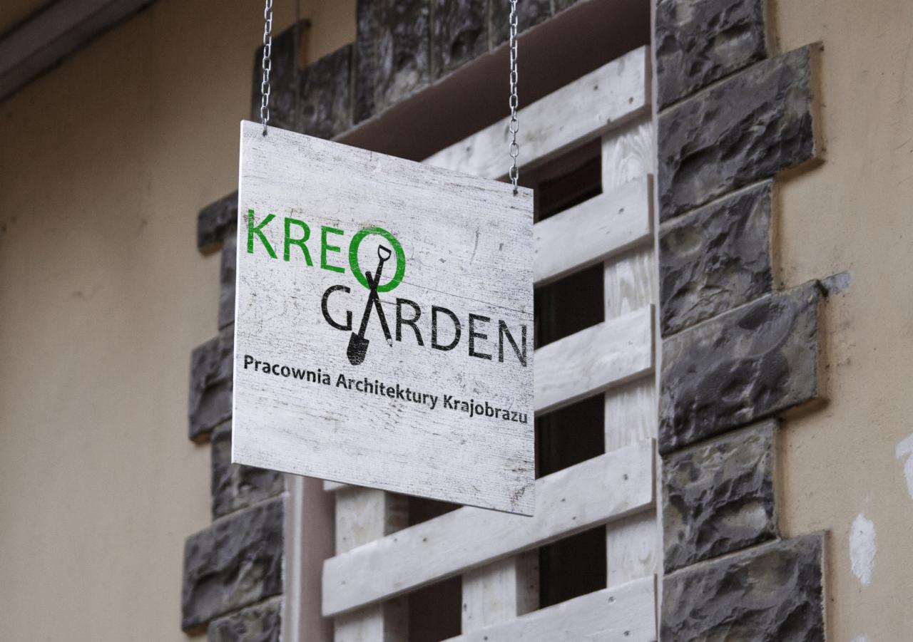 Szyld reklamowy z napisem 'KREO GARDEN Pracownia Architektury Krajobrazu' zawieszony na łańcuszkach przed budynkiem z elementami dekoracyjnymi.
