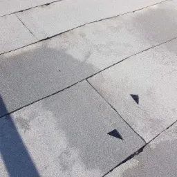 Szary dach pokryty papą z widocznymi łączeniami i łatami w kształcie trójkątów, częściowo mokry.