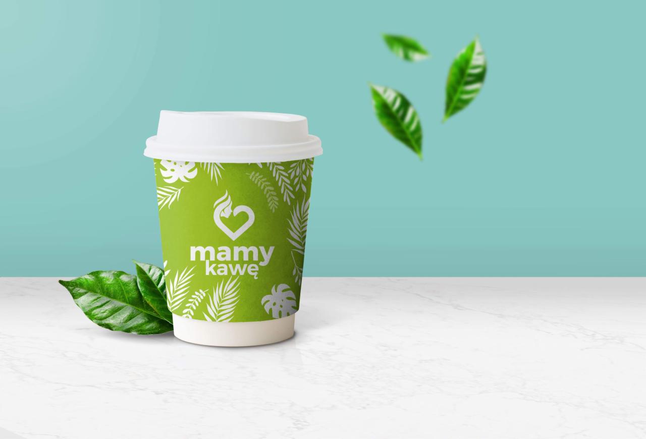Zielony kubek na kawę z logo 'mamy kawę' i motywem liści, otoczony świeżymi liśćmi kawy na białym blacie i tle w kolorze miętowym.