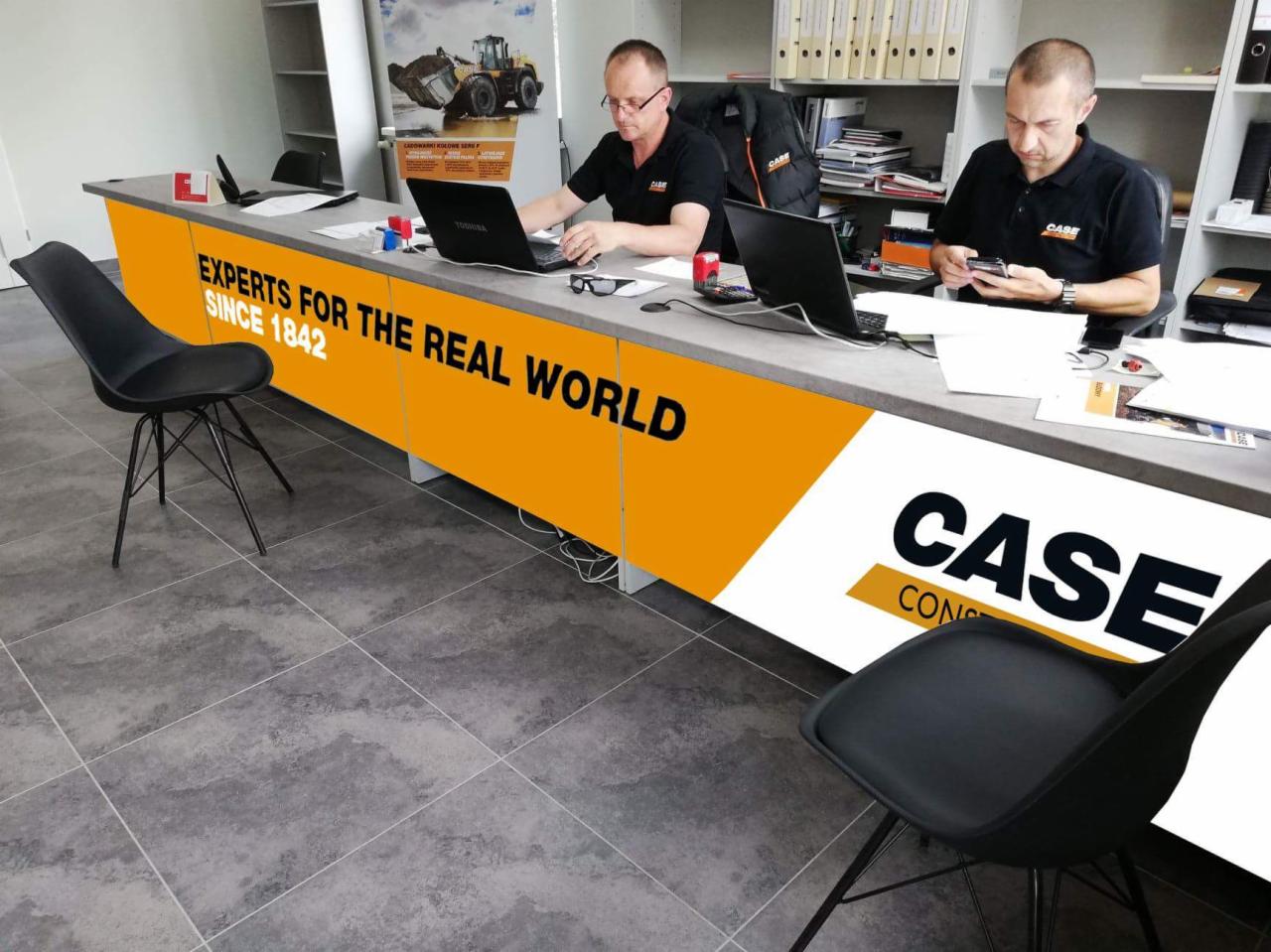 Biuro firmy CASE Construction Equipment: Dwóch mężczyzn w czarnych koszulkach z logo firmy pracuje przy biurku z laptopami i telefonem, na tle półki z segregatorami i plakatu z maszynami budowlanymi.