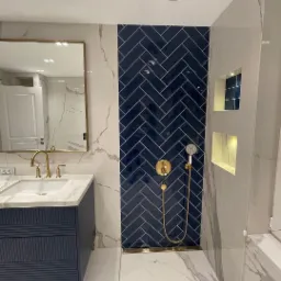 Nowoczesna łazienka z niebieską ścianą z płytek ułożonych w jodełkę, złotą armaturą i marmurowymi płytkami na ścianach i podłodze.