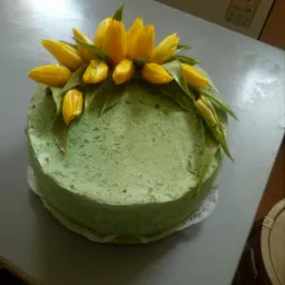 Okrągły tort z zielonym kremem, udekorowany żółtymi tulipanami i zielonymi liśćmi, umieszczony na białej koronkowej podkładce, na szarym stole. W tle lodówka z etykietą energetyczną.