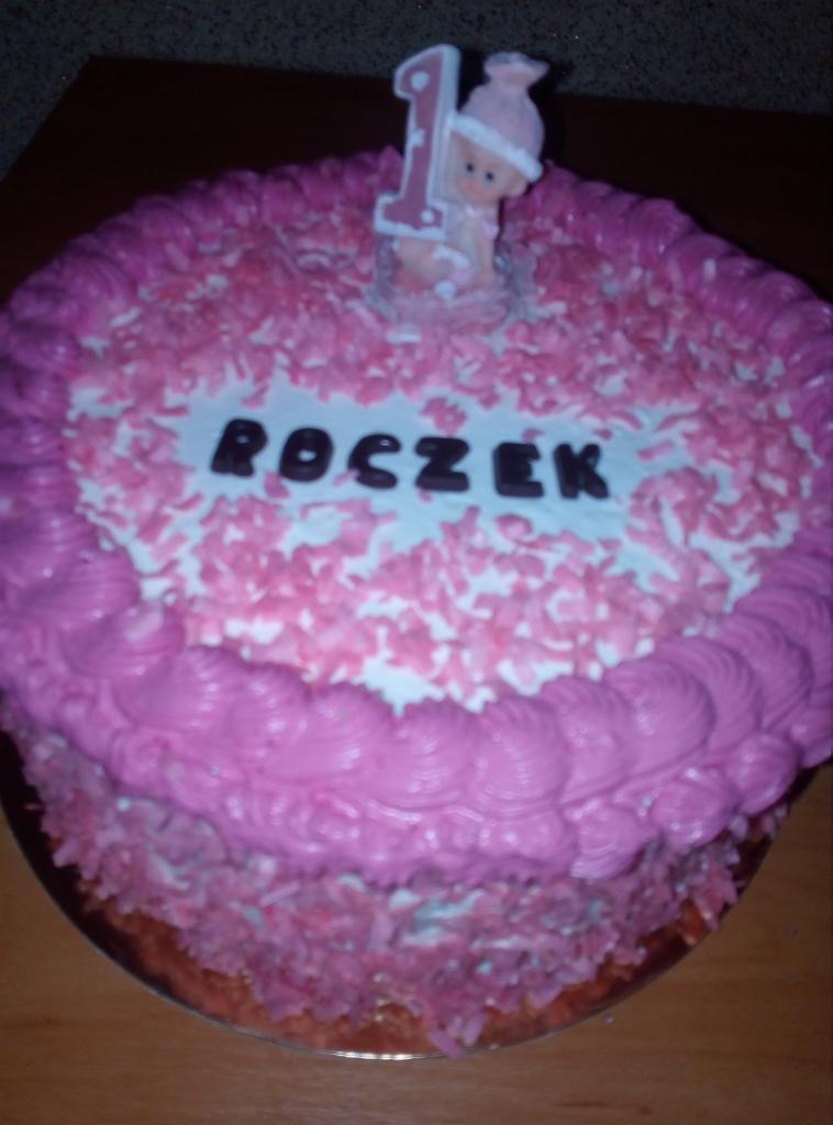Tort na roczek z różowym kremem, posypką i świeczką w kształcie cyfry 1 z figurką dziecka.