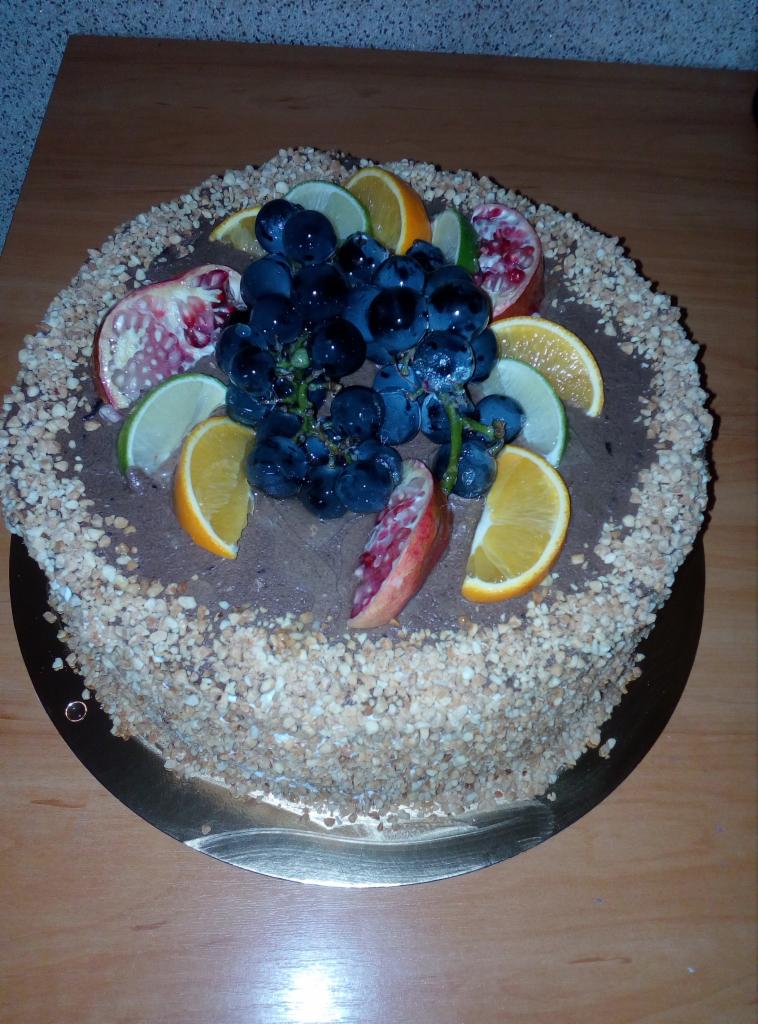 Okrągły tort z kremem czekoladowym, obsypany posiekanymi orzechami i udekorowany świeżymi owocami: winogronami, pomarańczami, limonkami i granatem.