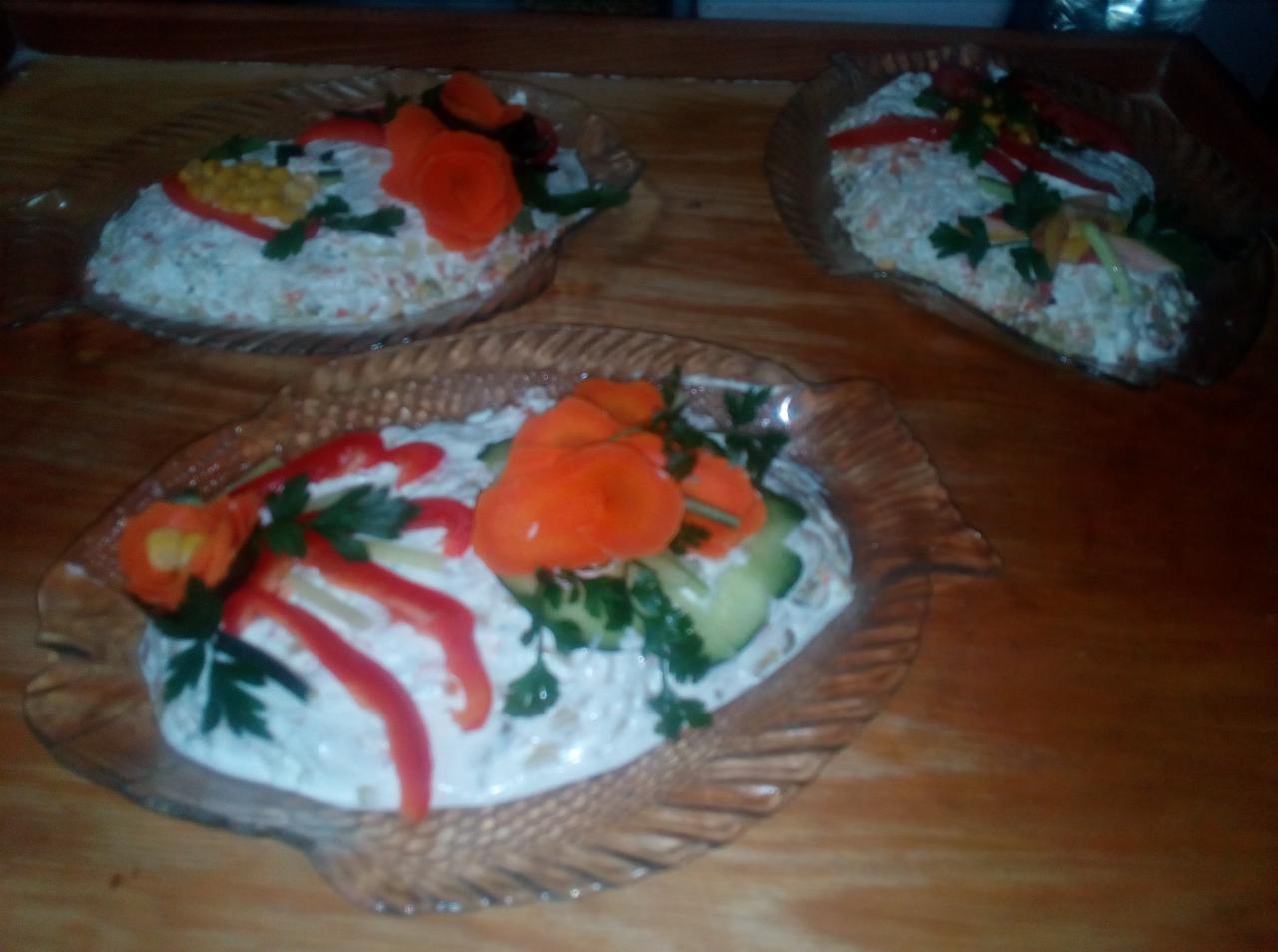 Trzy porcje sałatki jarzynowej ułożone w szklanych miseczkach w kształcie ryb, udekorowane różyczkami z marchewki, paskami czerwonej papryki i natką pietruszki na drewnianym stole.