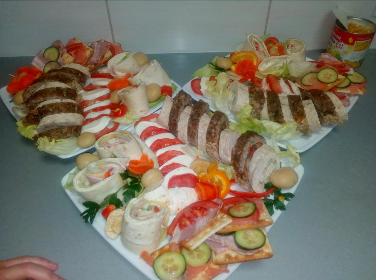 Dekoracyjne półmiski z roladkami z tortilli, plastrami mięsa, mozzarelli z pomidorami i krakersami z wędliną i ogórkiem, uzupełnione sałatą i warzywami, obok puszka kukurydzy.