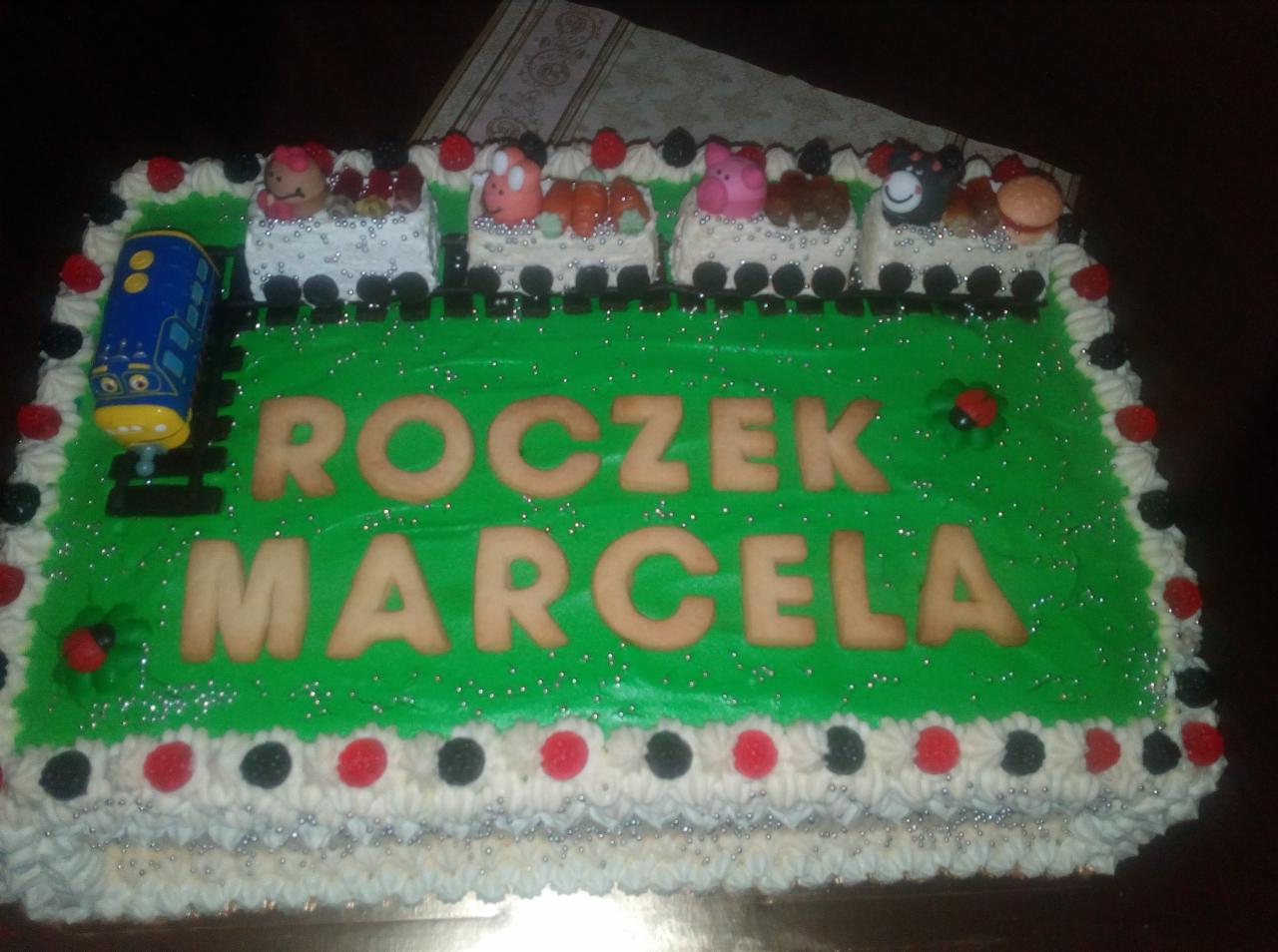 Prostokątny tort z zielonym lukrem, dekorowany ciastkowymi literami 'ROCZEK MARCELA',  cukrowym pociągiem i wagonikami ze zwierzątkami, obrzeża z kremowych rozetek i czerwono-czarnych koralików.