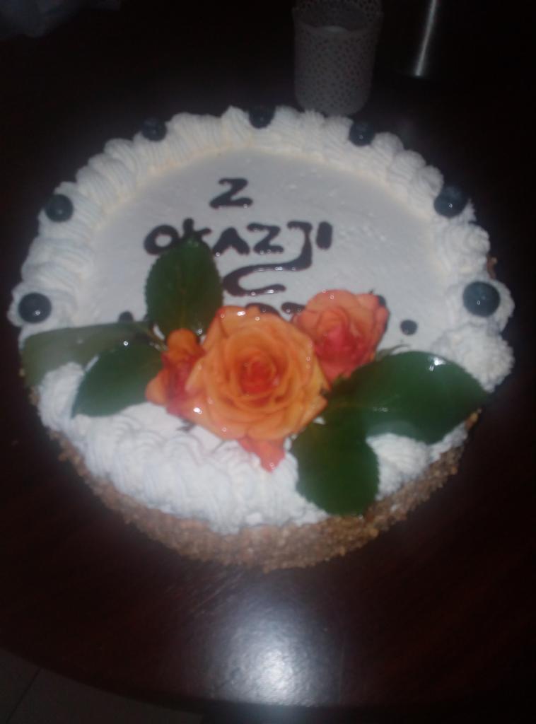 Tort urodzinowy z kremem, dekorowany różami i borówkami, z napisem 'Z Okazji', wykonany przez cukiernię z Międzyborza.