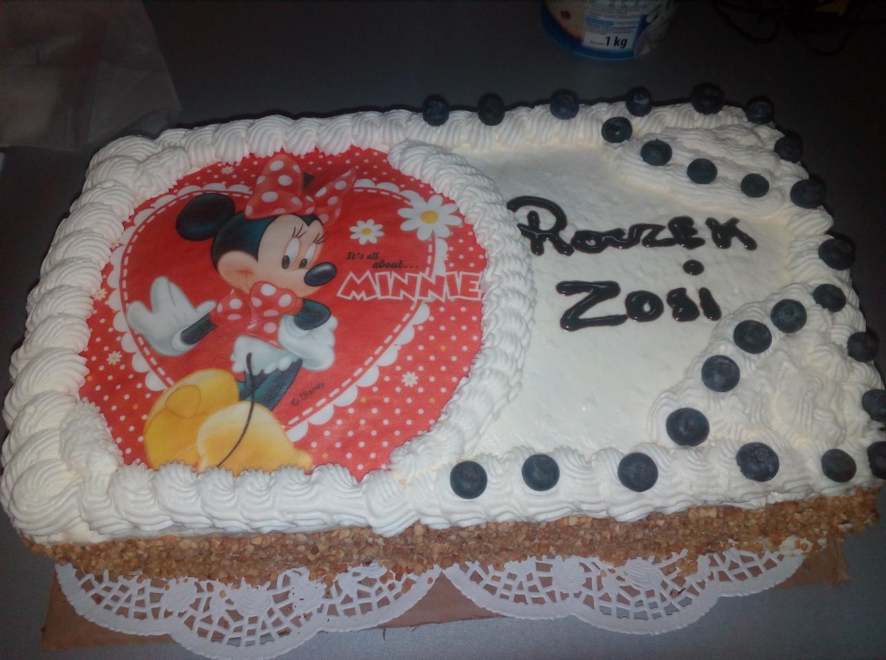 Prostokątny tort urodzinowy udekorowany kremem, borówkami i jadalnym nadrukiem z Myszką Minnie w czerwonym ubranku, na spodzie widoczna warstwa z orzechów i papierowa serwetka.
