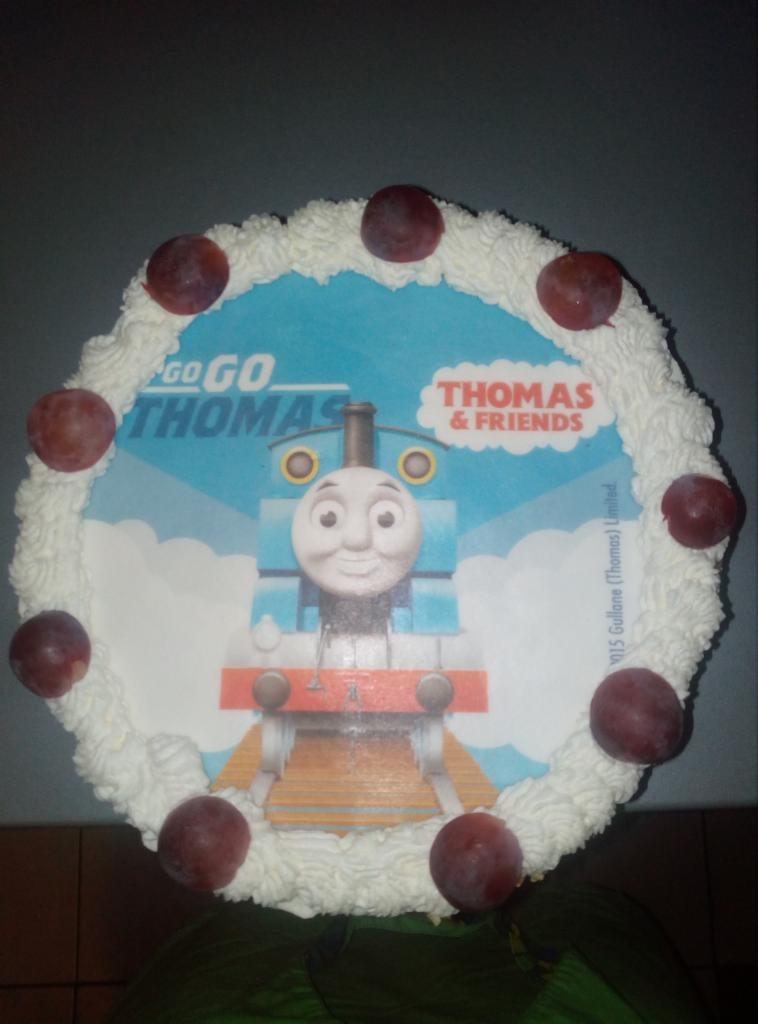 Okrągły tort z kremem, dekorowany winogronami i jadalnym nadrukiem z wizerunkiem lokomotywy Tomek i Przyjaciele.