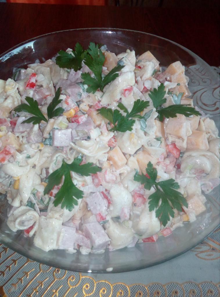 Szklana misa wypełniona sałatką makaronową z szynką, serem, papryką i kukurydzą, udekorowana świeżą natką pietruszki na ozdobnym obrusie.