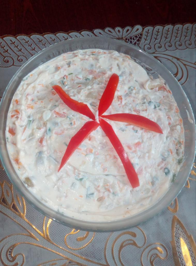 Sałatka jarzynowa w szklanej misce, udekorowana pięcioma paskami czerwonej papryki ułożonymi w kształt kwiatu, na wzorzystym obrusie.