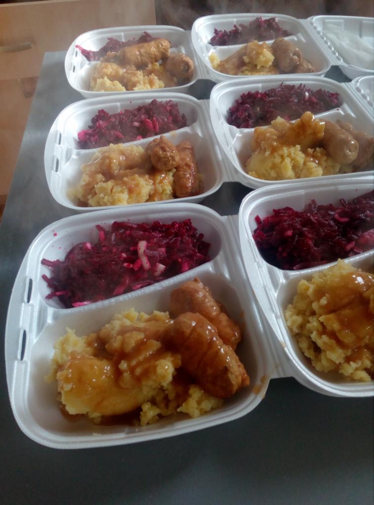 Porcje obiadowe w styropianowych pojemnikach: puree ziemniaczane z sosem, buraczki i dwie parówki, przygotowane do transportu.