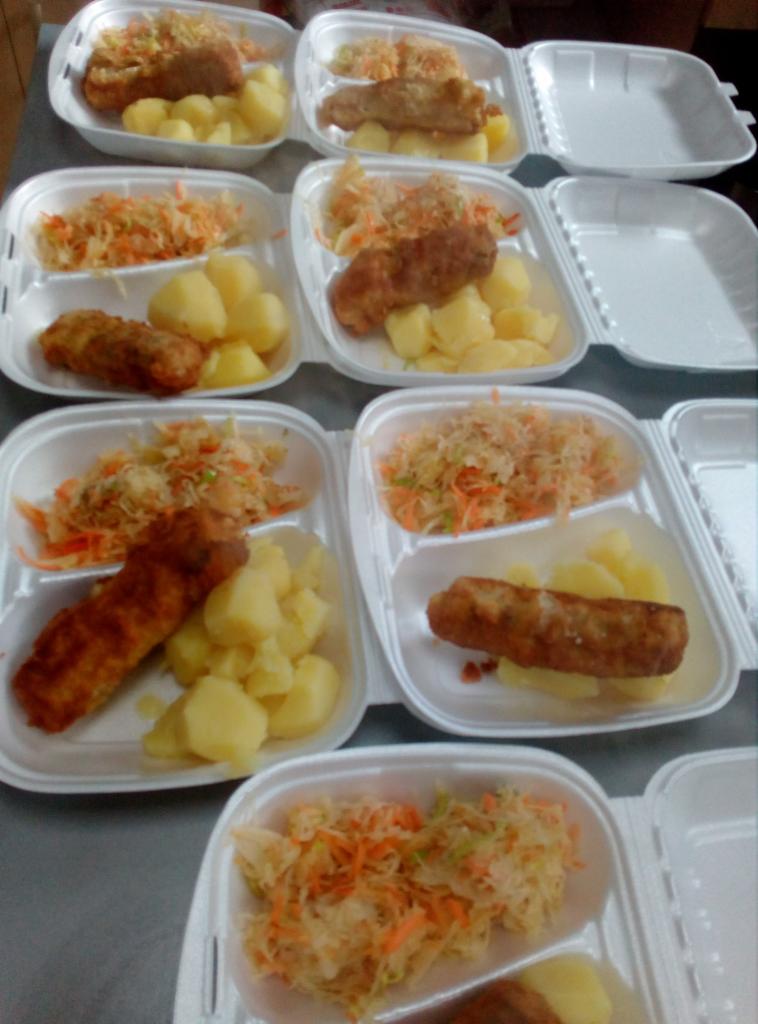Dania obiadowe w białych, styropianowych pojemnikach: kotlet, ziemniaki i surówka z kapusty i marchewki, ułożone rzędami, gotowe do transportu.