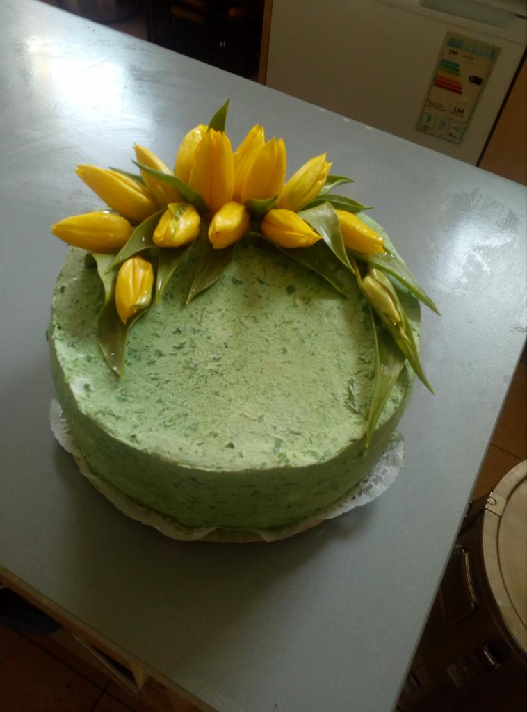 Okrągły tort z zielonym kremem, udekorowany żółtymi tulipanami i zielonymi liśćmi, umieszczony na białej koronkowej podkładce, na szarym stole. W tle lodówka z etykietą energetyczną.