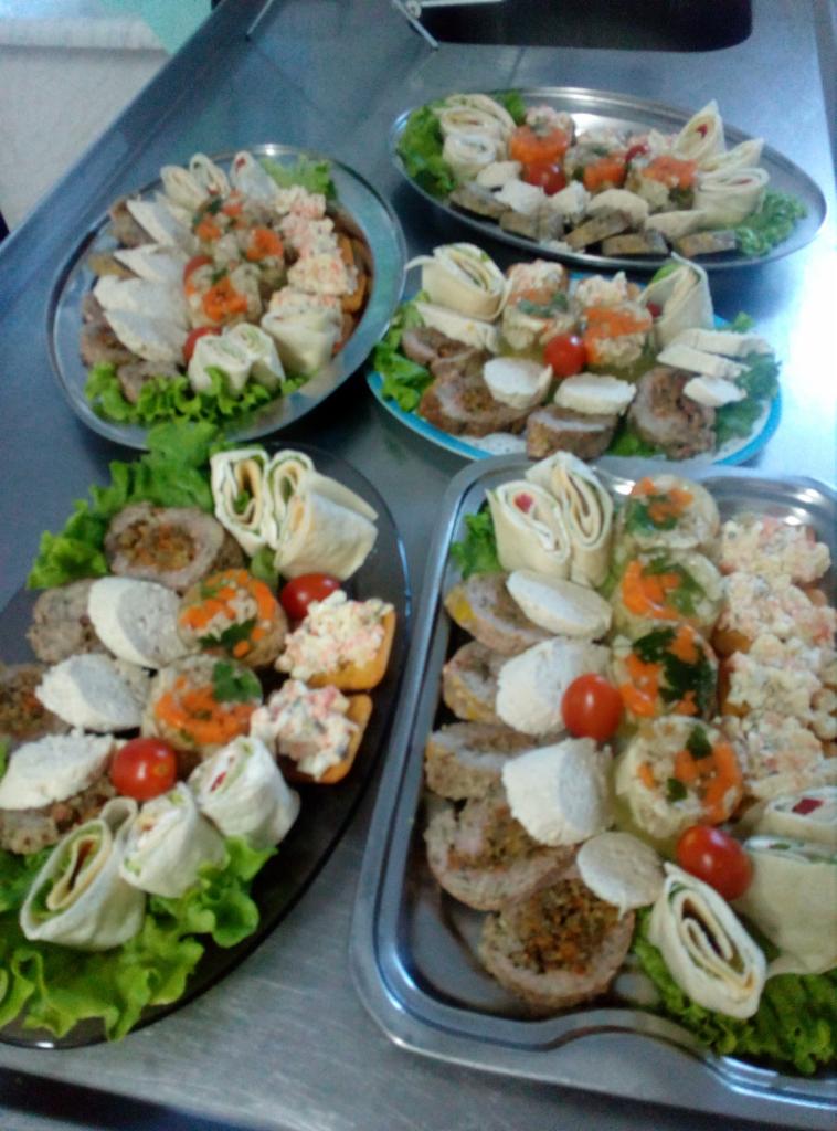 Różnorodne przekąski na czterech metalowych tacach: roladki z tortilli, koreczki, mięsne roladki z białym serem, galaretki z warzywami, sałatki, udekorowane listkami sałaty i pomidorkami koktajlowymi.