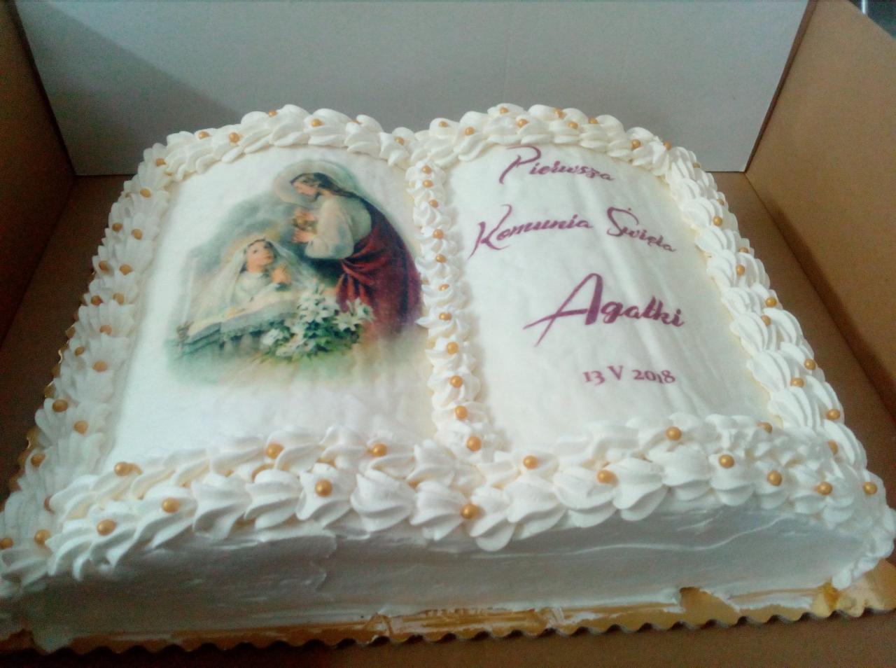 Tort w kształcie otwartej księgi, dekorowany białym kremem i złotymi kuleczkami, z nadrukiem przedstawiającym Jezusa z dziewczynką oraz napisem 'Pierwsza Komunia Święta Agatki 13 V 2018'.