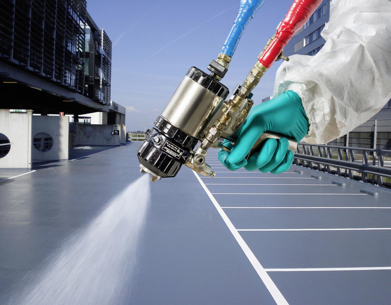 Aplikacja szarej powłoki na posadzce parkingu wielopoziomowego za pomocą pistoletu natryskowego; pracownik w kombinezonie ochronnym i zielonych rękawicach trzyma urządzenie, w tle budynek biurowy.