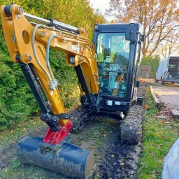 Żółto-czarna mini koparka SANY na gąsienicach w trakcie pracy w ogrodzie, widoczny wykopany wąski rów w ziemi, w tle zielony żywopłot i zaparkowany biały bus.