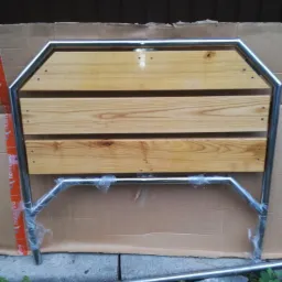 Konstrukcja z trzech drewnianych desek w metalowej ramie, zabezpieczona folią, oparta o karton na zewnątrz budynku.