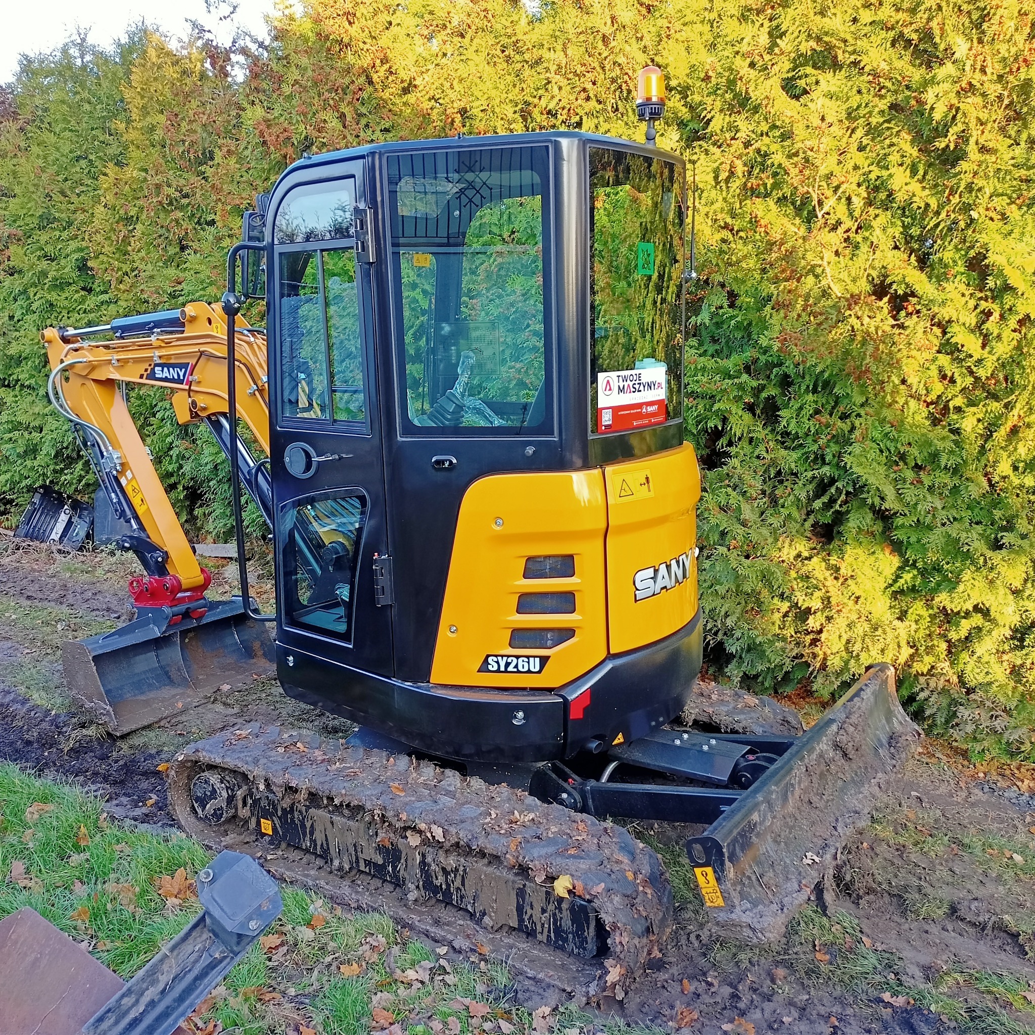 Żółto-czarna mini koparka SANY SY26U na gąsienicach, stojąca na ziemi z widocznymi śladami pracy, na tle zielonych krzewów.