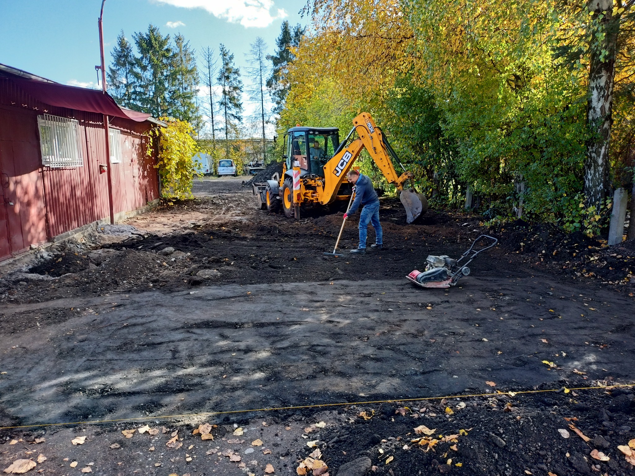 Żółta koparko-ładowarka JCB podczas wyrównywania terenu podłoża przed budynkiem, pracownik z grabiami, ubijarka w tle, jesienne drzewa w tle, częściowo przygotowany teren.