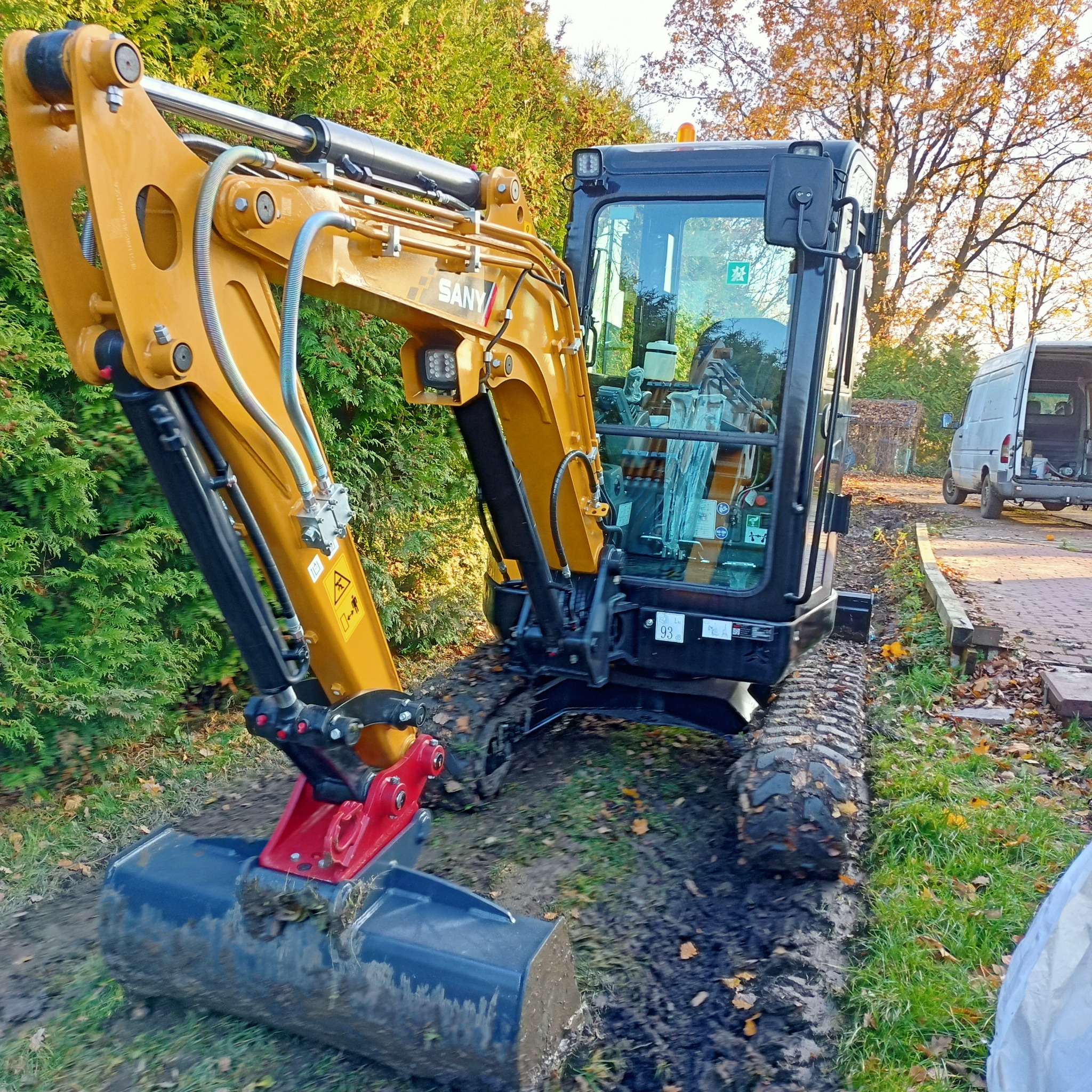 Żółto-czarna mini koparka SANY na gąsienicach w trakcie pracy w ogrodzie, widoczny wykopany wąski rów w ziemi, w tle zielony żywopłot i zaparkowany biały bus.
