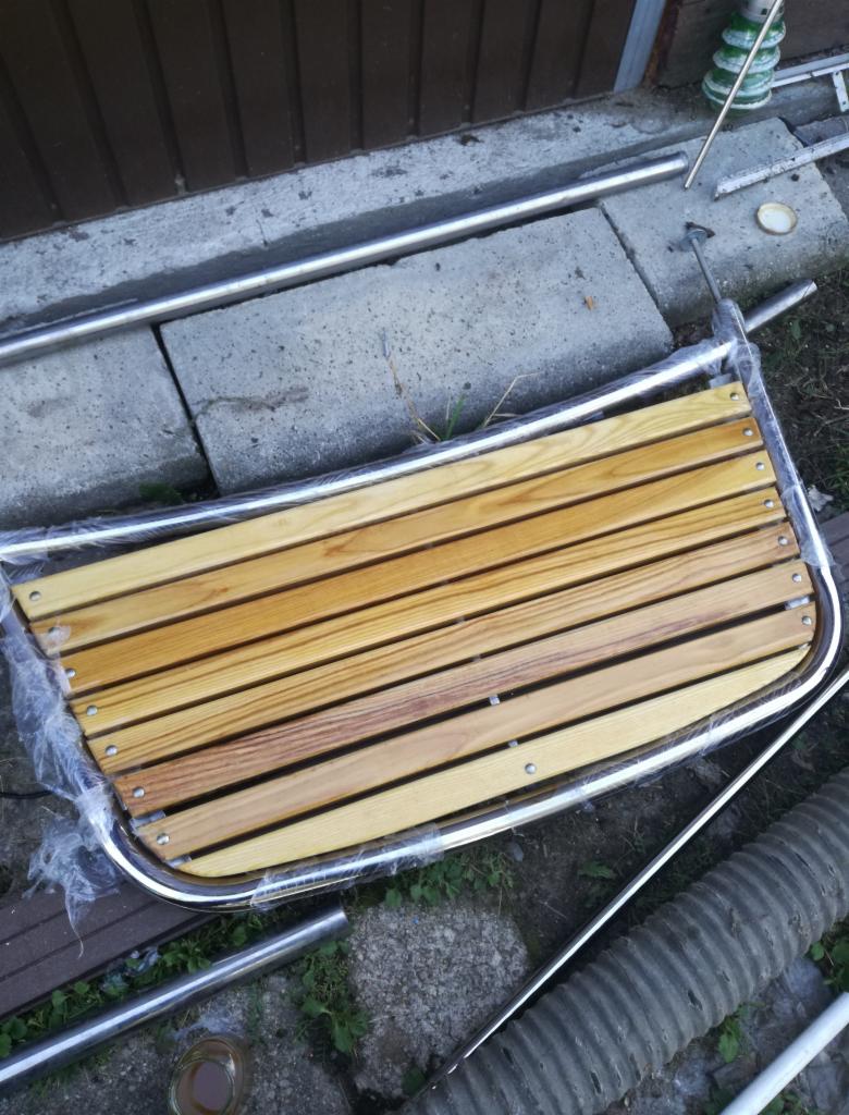 Element konstrukcyjny z drewnianych listew i chromowanej ramy, owinięty folią, leżący na ziemi obok betonowych płyt i rur.