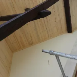 Drewniana boazeria na poddaszu z widocznymi belkami stropowymi w kolorze ciemnego brązu, ujęcie z dołu, widoczny fragment rusztowania.