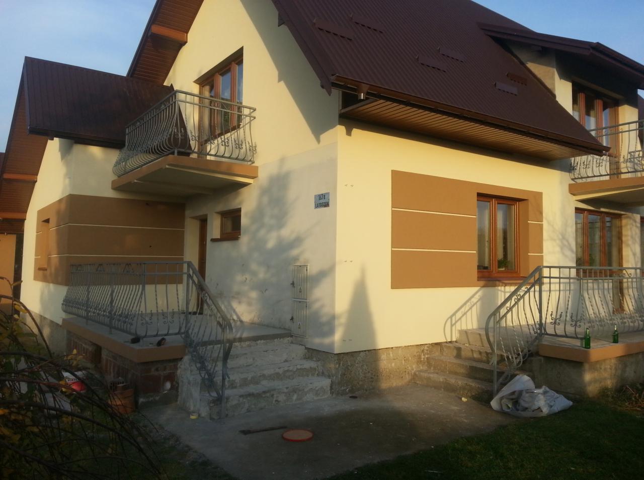 Dwukondygnacyjny dom z jasną elewacją i brązowymi akcentami, widoczne balkony z metalowymi balustradami, brązowy dach, schody wejściowe i fragment ogrodu.