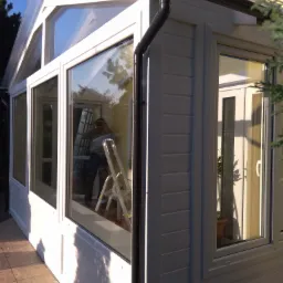 Biała, przeszklona konstrukcja dobudowana do domu, z widoczną rynną odprowadzającą wodę i odbiciem osoby robiącej zdjęcie na drabinie w szybie.