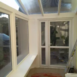 Wnętrze jasnej oranżerii z przeszklonym dachem, białymi ramami okiennymi i drzwiami, widoczna drabina oparta o żółtą ścianę i odbicie osoby robiącej zdjęcie w szybie.