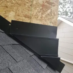 Czarny, metalowy element wykończeniowy dachu, zamontowany na krawędzi przy ścianie budynku, widoczne gwoździe mocujące i struktura papy dachowej.