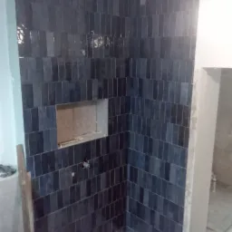 Ściana w trakcie wykańczania, pokryta ciemnymi, błyszczącymi płytkami ułożonymi pionowo, z wnęką na półkę i wystającymi elementami instalacji.