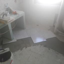 Raman construction fcb - Układanie jasnych, kwadratowych płytek podłogowych w łazience, widoczny fragment wanny w folii ochronnej, narzędzia glazurnicze i zapas płytek na prowizorycznym blacie z płyt gipsowo-kartonowych...