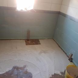 Raman construction fcb - Świeżo nałożona warstwa kleju do płytek na podłodze w trakcie prac w łazience, widoczne rury wystające z podłoża i częściowo wykończone ściany z szarymi płytkami.