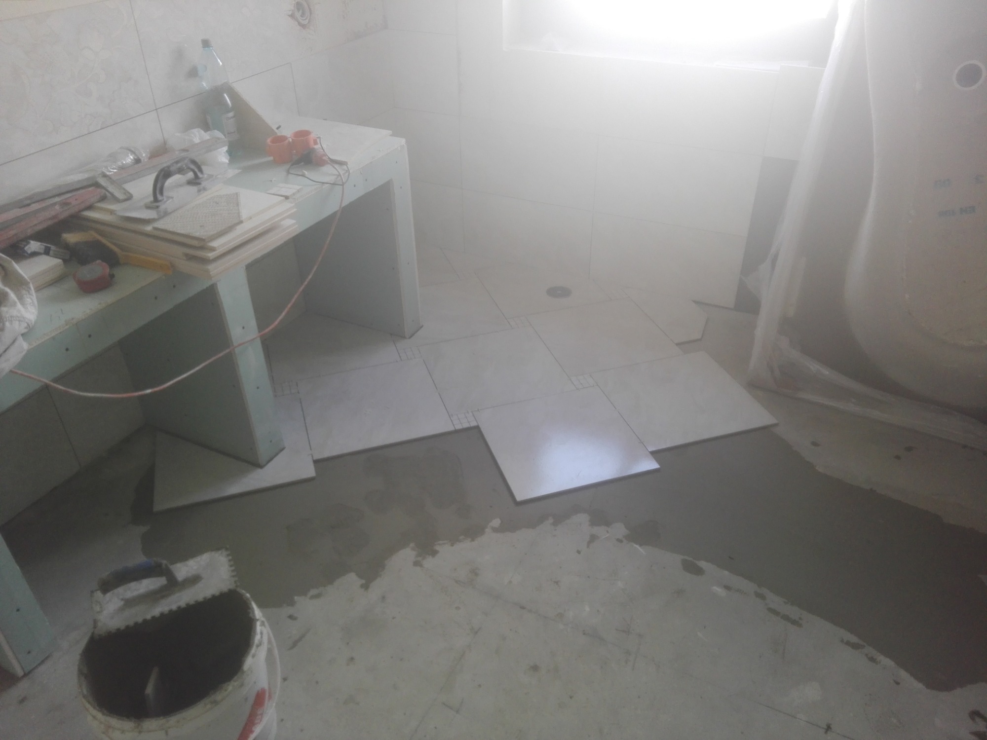 Układanie jasnych, kwadratowych płytek podłogowych w łazience, widoczny fragment wanny w folii ochronnej, narzędzia glazurnicze i zapas płytek na prowizorycznym blacie z płyt gipsowo-kartonowych...