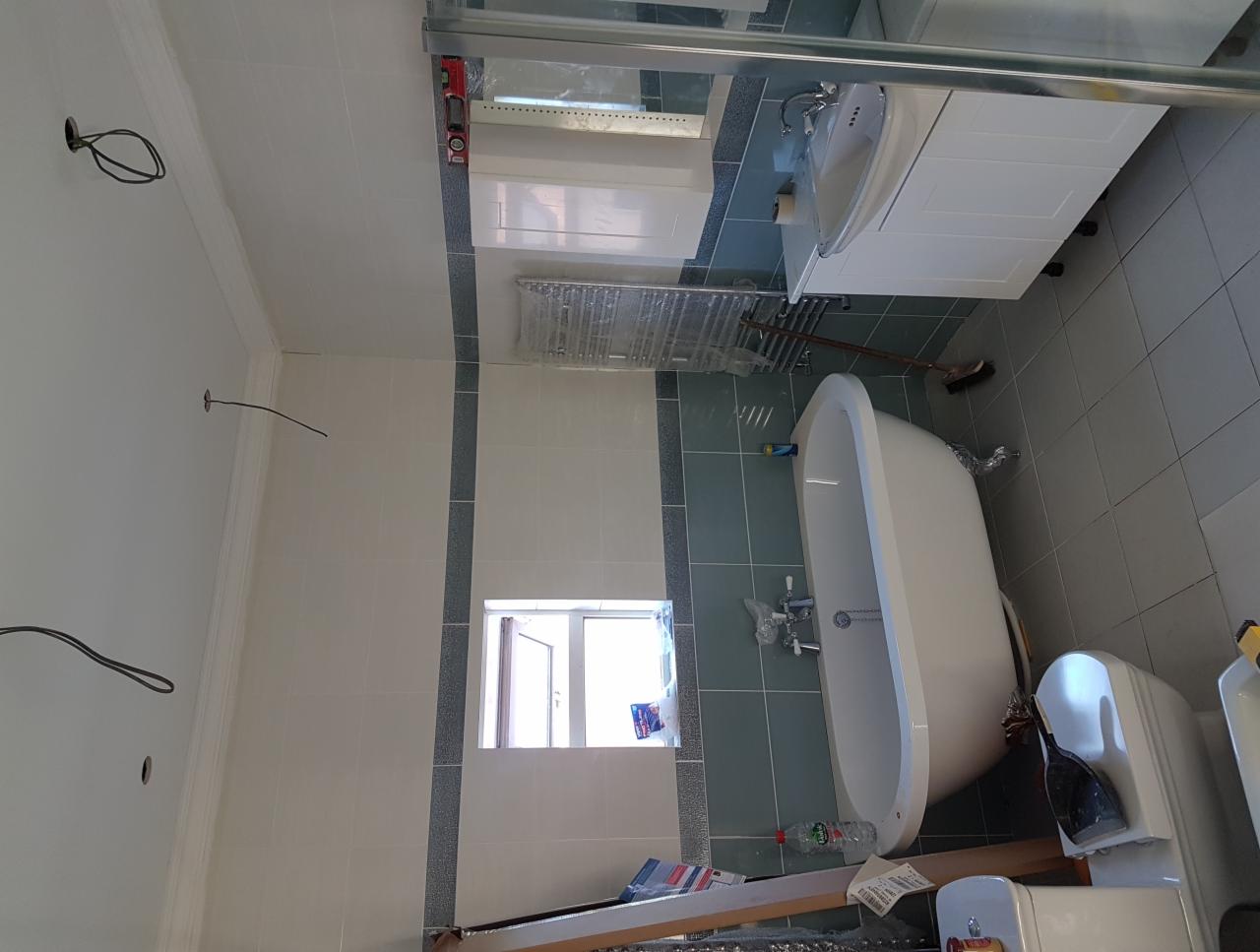 Wnętrze łazienki w trakcie prac wykończeniowych, widoczna wanna wolnostojąca, umywalka z szafką, grzejnik drabinkowy, płytki na ścianach i podłodze, okno oraz wystające przewody elektryczne ze ściany.