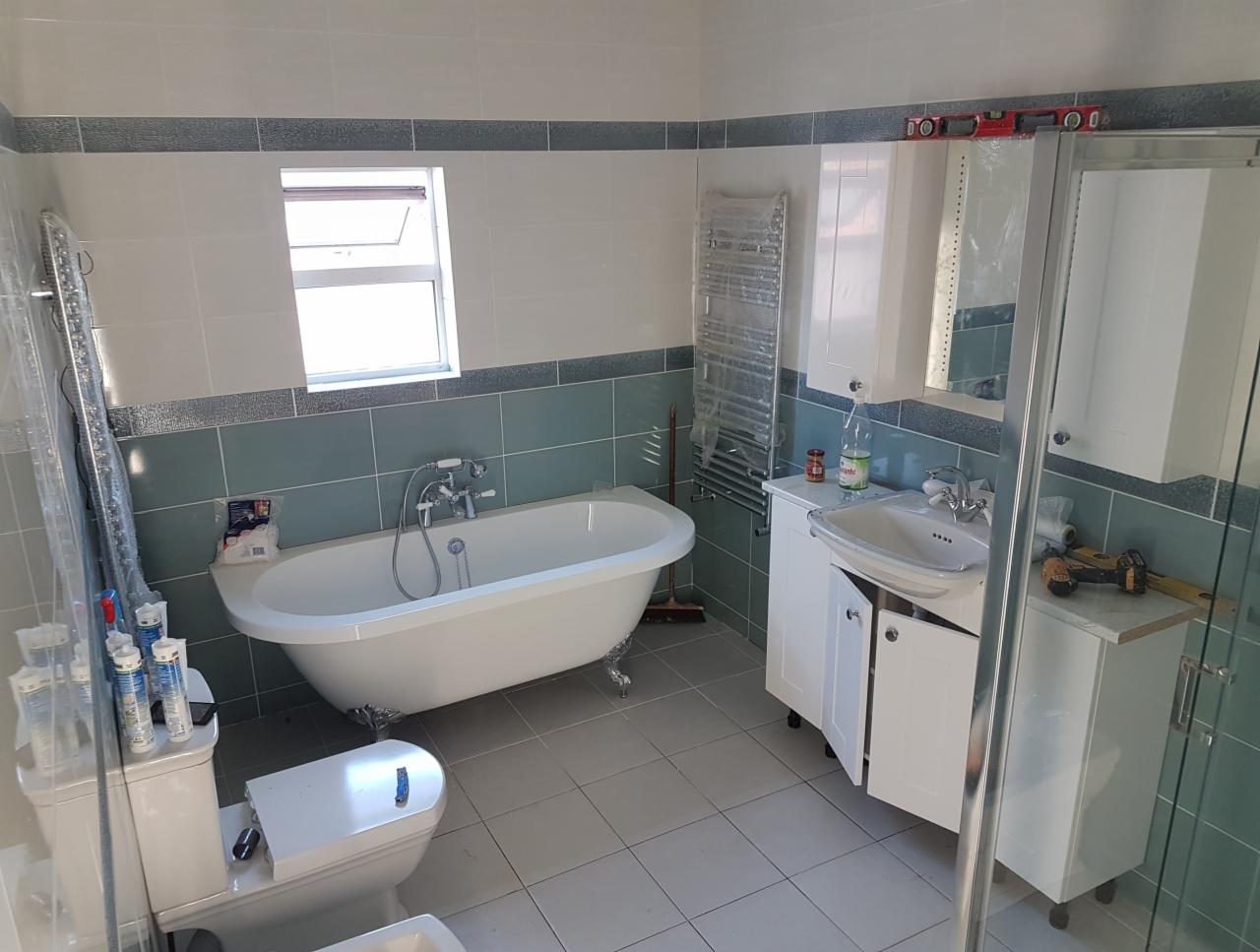 Wykończona łazienka z wolnostojącą wanną na lwich nóżkach, białą umywalką z szafką, grzejnikiem drabinkowym w folii ochronnej, kabiną prysznicową i poziomnicą na szafce.