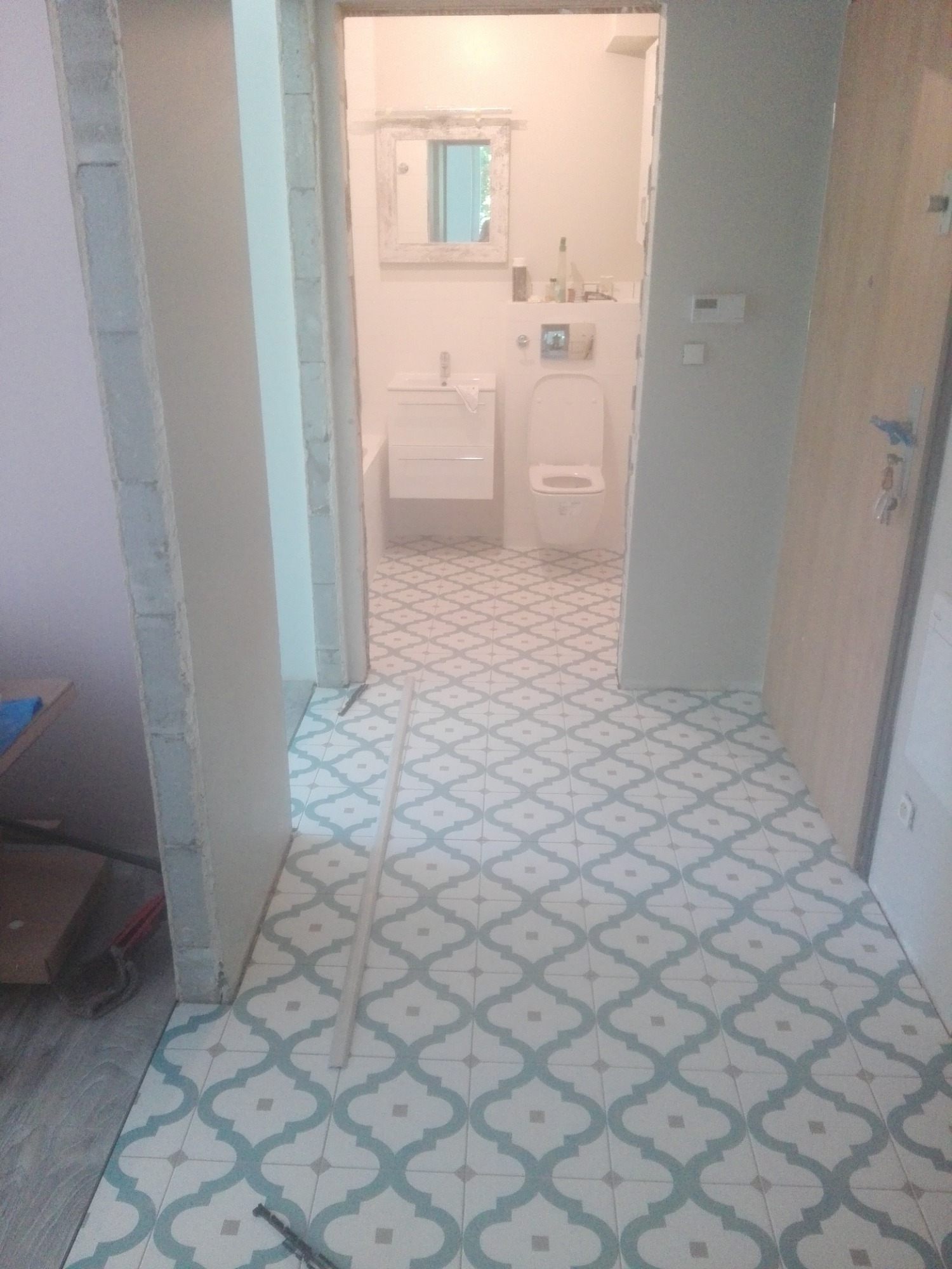 Ułożona podłoga z wzorzystych płytek w odcieniach bieli i błękitu, widok na częściowo wykończoną łazienkę z białą armaturą i lustrem w ozdobnej ramie, surowy betonowy fragment ściany po lewej stronie.