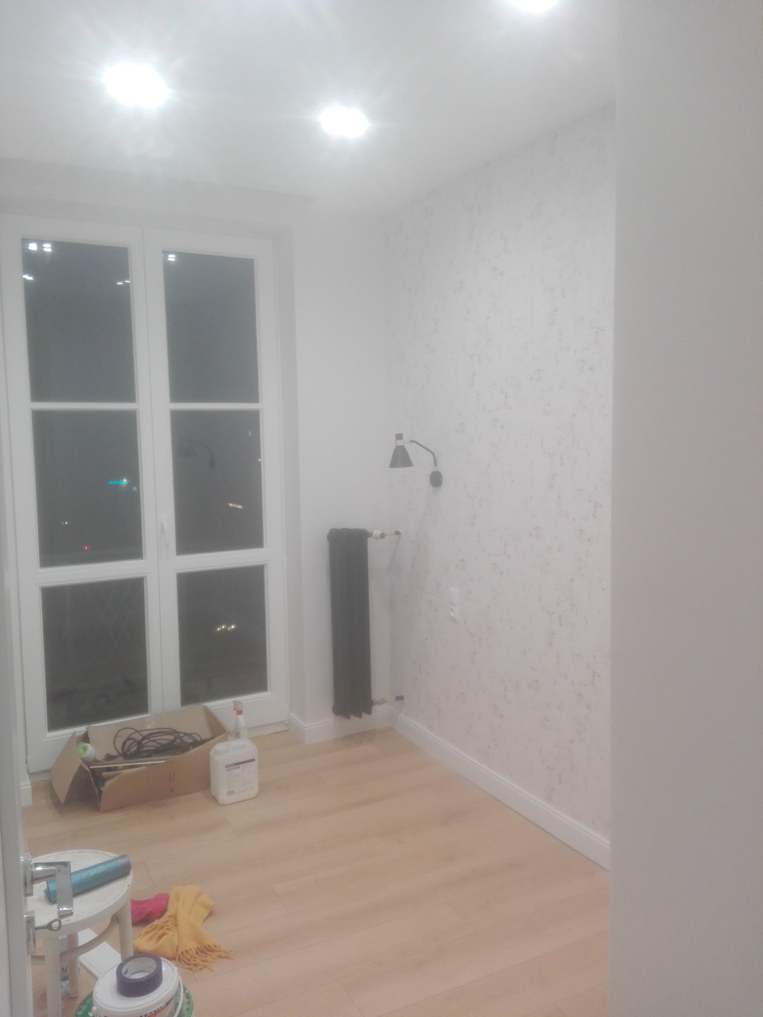 Pokój w trakcie remontu: ściana z tapetą w odcieniach bieli i beżu, jasne panele podłogowe, białe okno z widokiem na miasto, czarny grzejnik i lampka ścienna, karton z kablami i pojemnik z płynem...