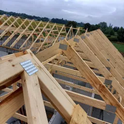 Konstrukcja drewnianego dachu z metalowymi łącznikami, widok z góry na tle pochmurnego nieba i zieleni.