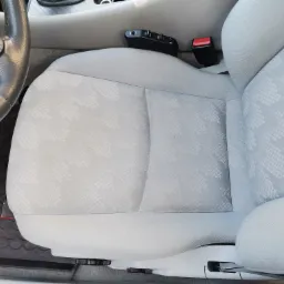 Jasnoszare, tekstylne siedzenie samochodowe z subtelnym wzorem, widoczny fragment kierownicy i panel regulacji fotela.