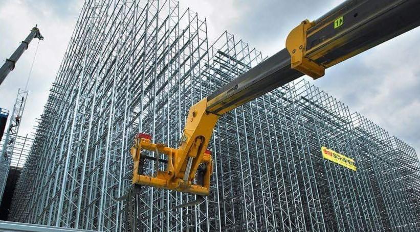 Montaż modułowej konstrukcji z metalowych profili na placu budowy, widoczny podnośnik koszowy i fragment dźwigu w tle.