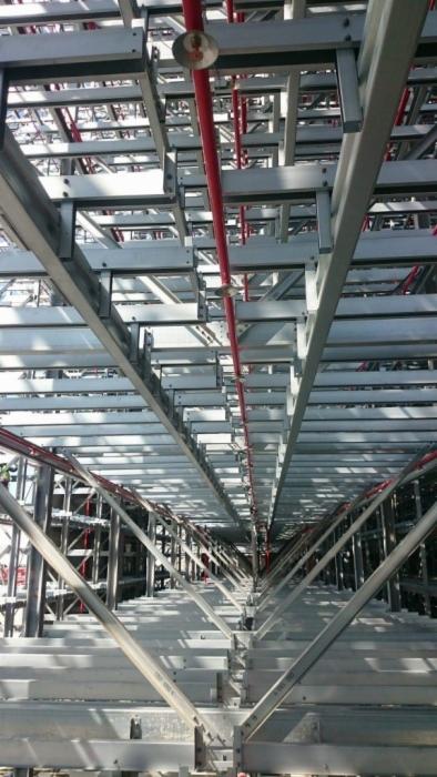 Wnętrze magazynu z widocznymi stalowymi regałami, czerwonym systemem rur przeciwpożarowych pod sufitem i perspektywą w głąb konstrukcji.