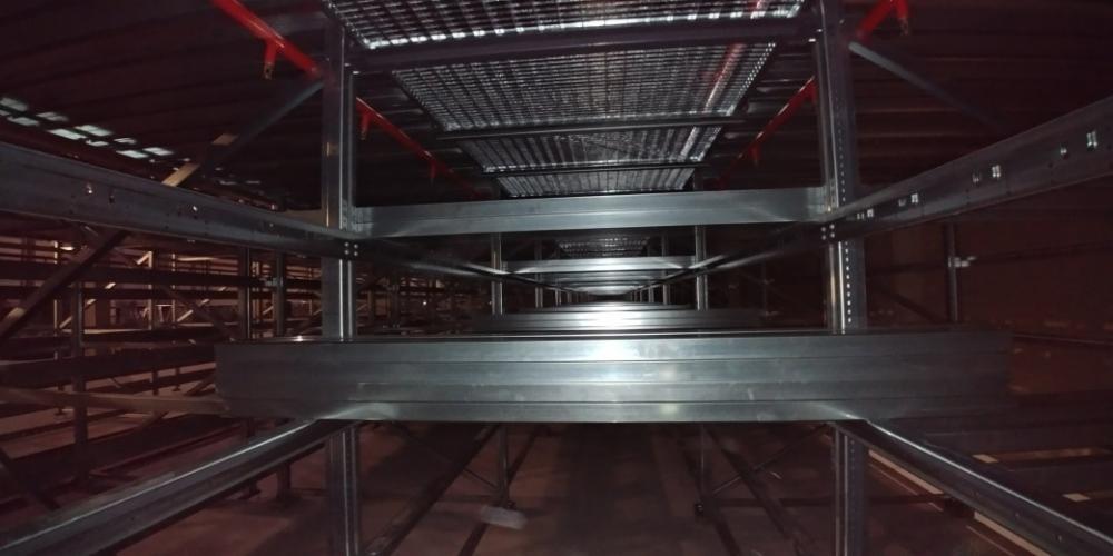 Wnętrze magazynu z zamontowanymi, wielopoziomowymi regałami z metalową kratką, widok od dołu wzdłuż korytarza między regałami.