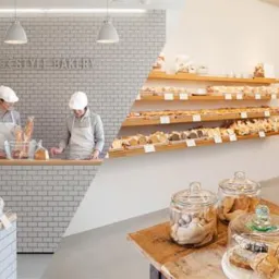 Jasne wnętrze cukierni z ekspozycją pieczywa na drewnianych półkach i pracownikami za ladą wyłożoną białymi cegiełkami, widoczne słoiki z ciastkami na drewnianym stole.