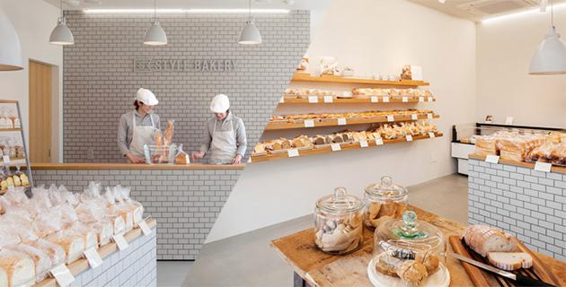 Jasne wnętrze cukierni z ekspozycją pieczywa na drewnianych półkach i pracownikami za ladą wyłożoną białymi cegiełkami, widoczne słoiki z ciastkami na drewnianym stole.