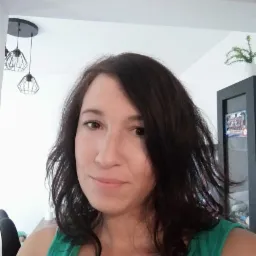 Portret kobiety z ciemnymi włosami, uśmiechającej się, w zielonej koszulce na tle jasnego wnętrza z lampami i szafką. Ujęcie z bliska, naturalne światło.