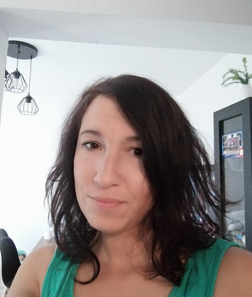 Portret kobiety z ciemnymi włosami, uśmiechającej się, w zielonej koszulce na tle jasnego wnętrza z lampami i szafką. Ujęcie z bliska, naturalne światło.